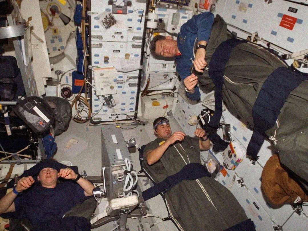 Three astronauts sleep in sleeping bags attached to the walls and ceiling inside a spacecraft or space station, floating due to zero gravity. The area is filled with equipment, wires, and storage compartments.