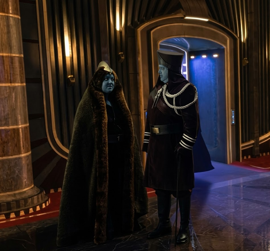 Two people with blue skin and dark clothing, including elaborate coats and hats, stand in a luxurious, futuristic hallway with metallic walls, golden accents, and a blue-lit doorway.