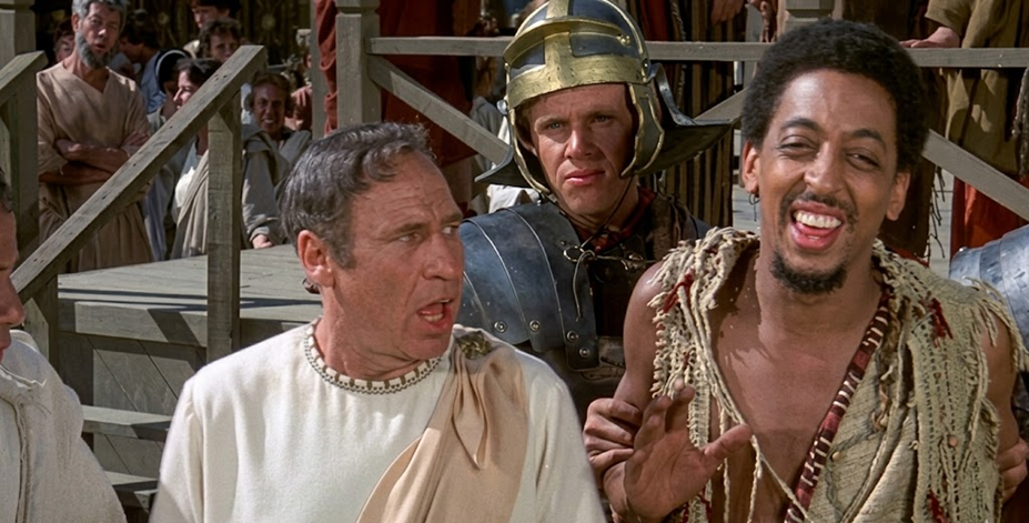 Three men stand in the foreground; one wears a white toga, another is dressed as a Roman soldier with a helmet, and the third wears rough, tattered clothing and is laughing. People and wooden structures are visible in the background.