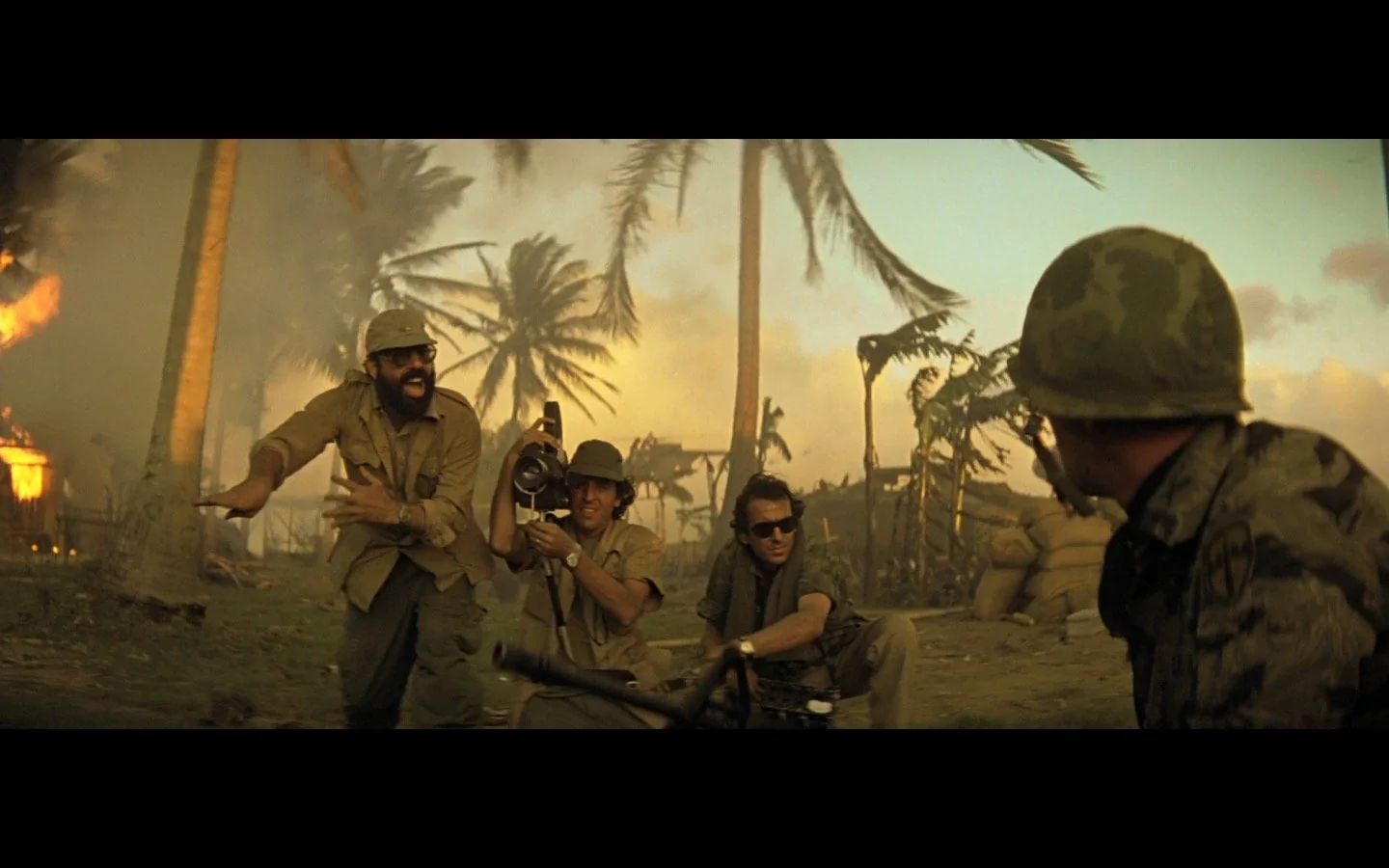 Three soldiers crouch and aim their weapons amid chaos, smoke, and swaying palm trees during a battle at sunset. Another soldier stands in the foreground, partially visible. The scene is intense and dramatic.