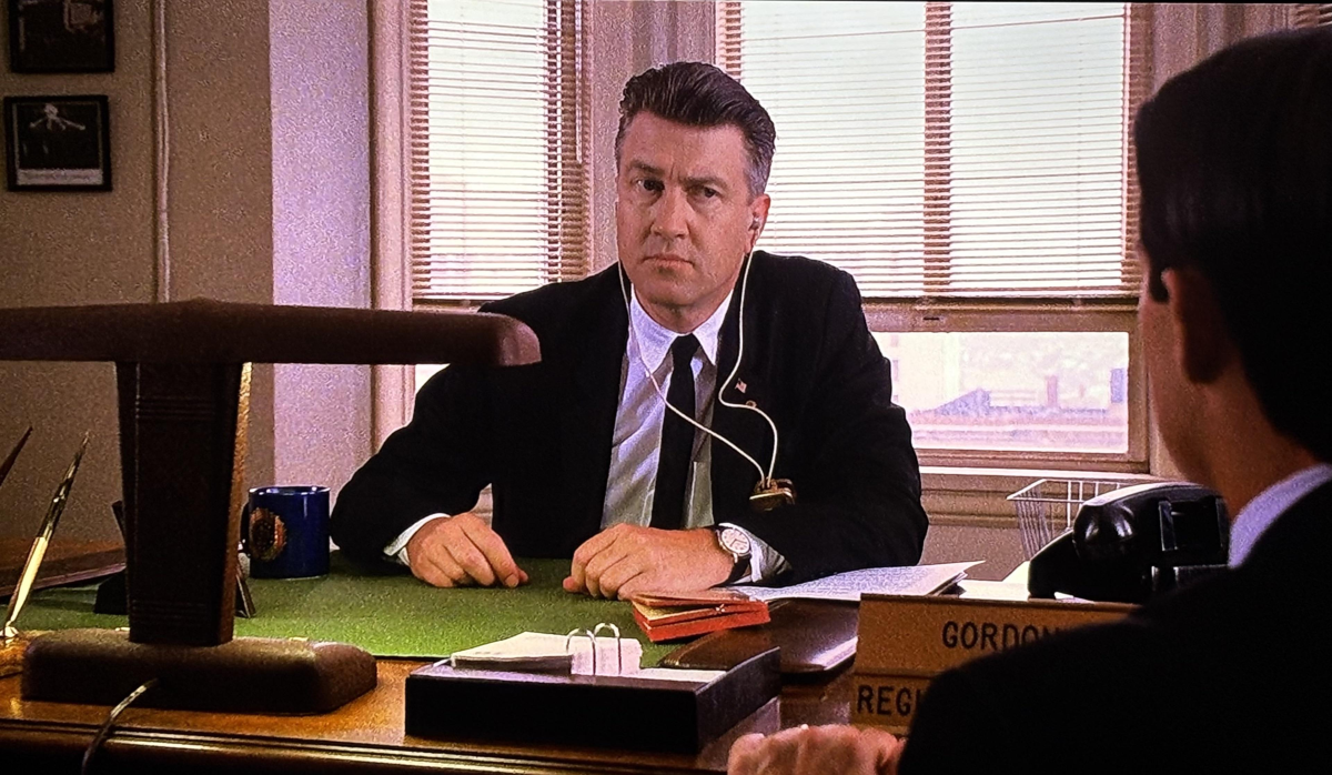 A man in a black suit with an earpiece sits at a desk, looking serious. There are papers, a mug, and a nameplate on the desk; venetian blinds cover the window behind him. Another person is partially visible in the foreground.
