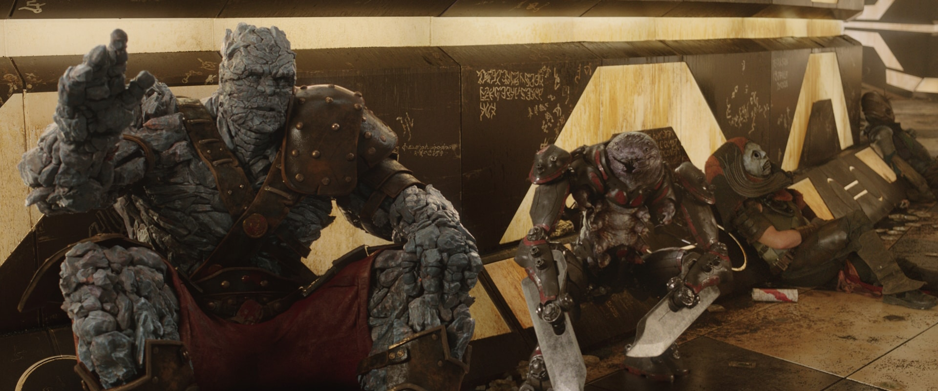 Three armored alien warriors with rocky and reptilian features sit against a futuristic, graffiti-covered wall, looking tired or defeated, in a sci-fi movie scene.