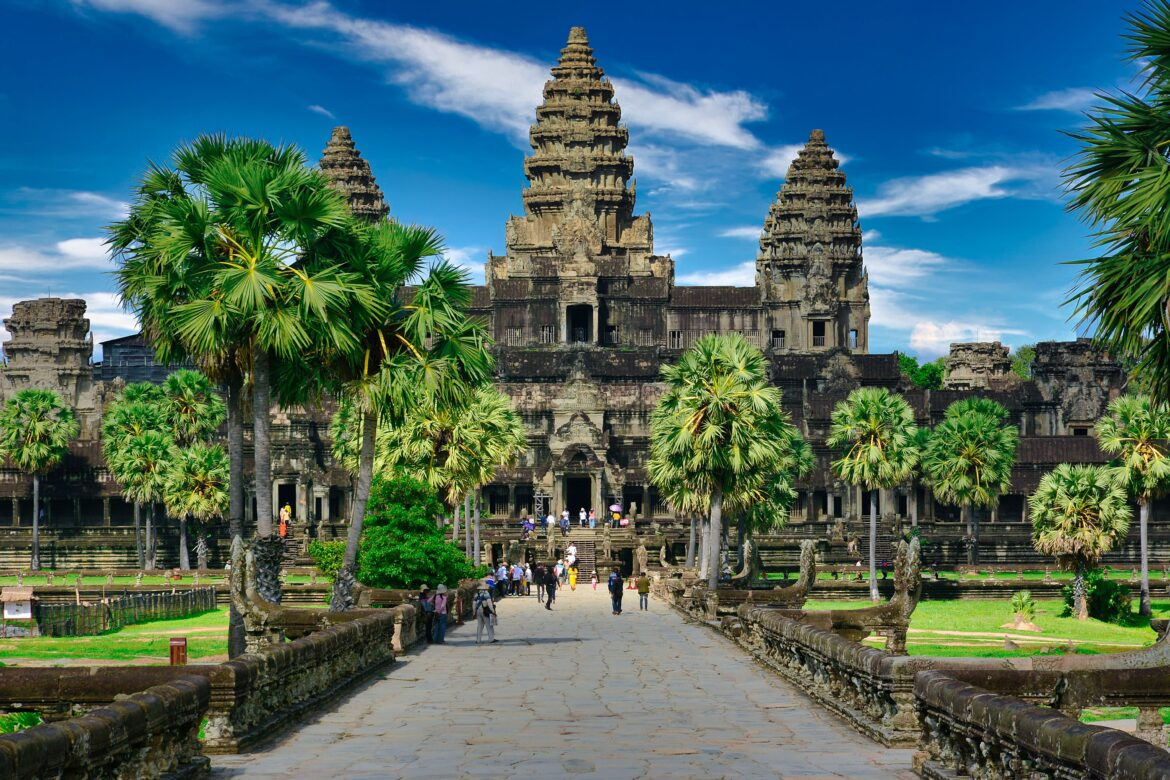 Angkor Wat temple in Cambodia, viewed from the stone walkway entrance, surrounded by palm trees, with tourists walking toward the ancient, intricately carved stone structure under a bright blue sky.