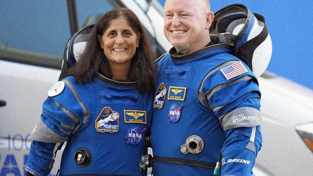 Two astronauts stand side by side smiling, wearing blue NASA spacesuits with patches and the U.S. flag. The background is blurred but suggests a space or aerospace setting.
