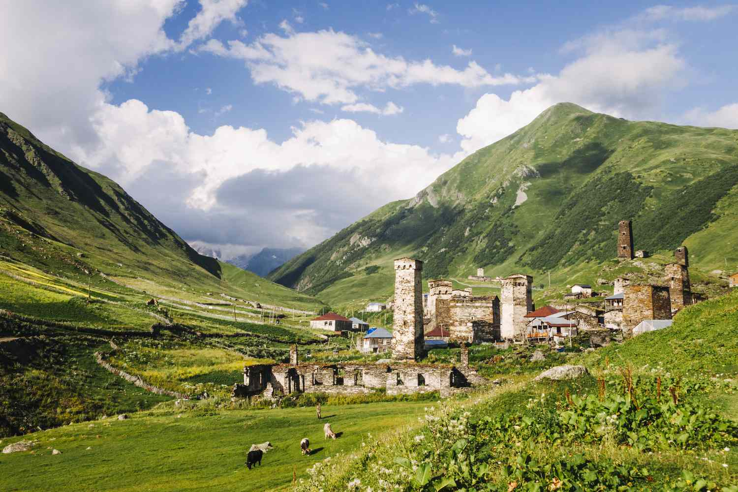 A lush green valley with grazing animals, stone towers, and village houses nestled between rolling mountains under a partly cloudy blue sky.