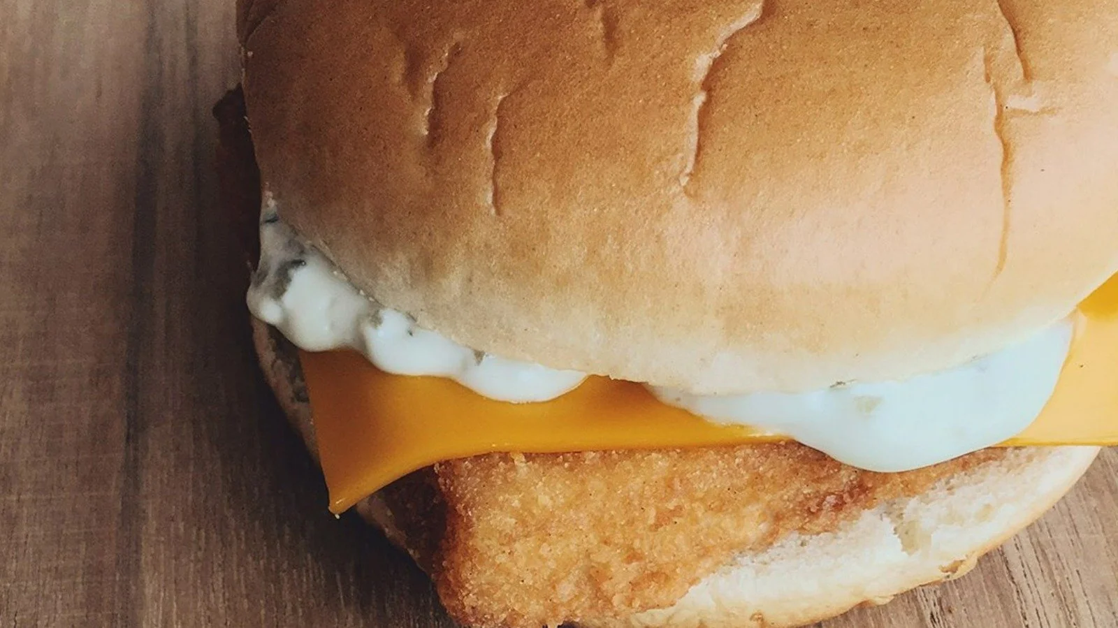 A close-up of a fish sandwich with a golden fried fish fillet, a slice of cheddar cheese, and tartar sauce in a soft hamburger bun, on a wooden surface.