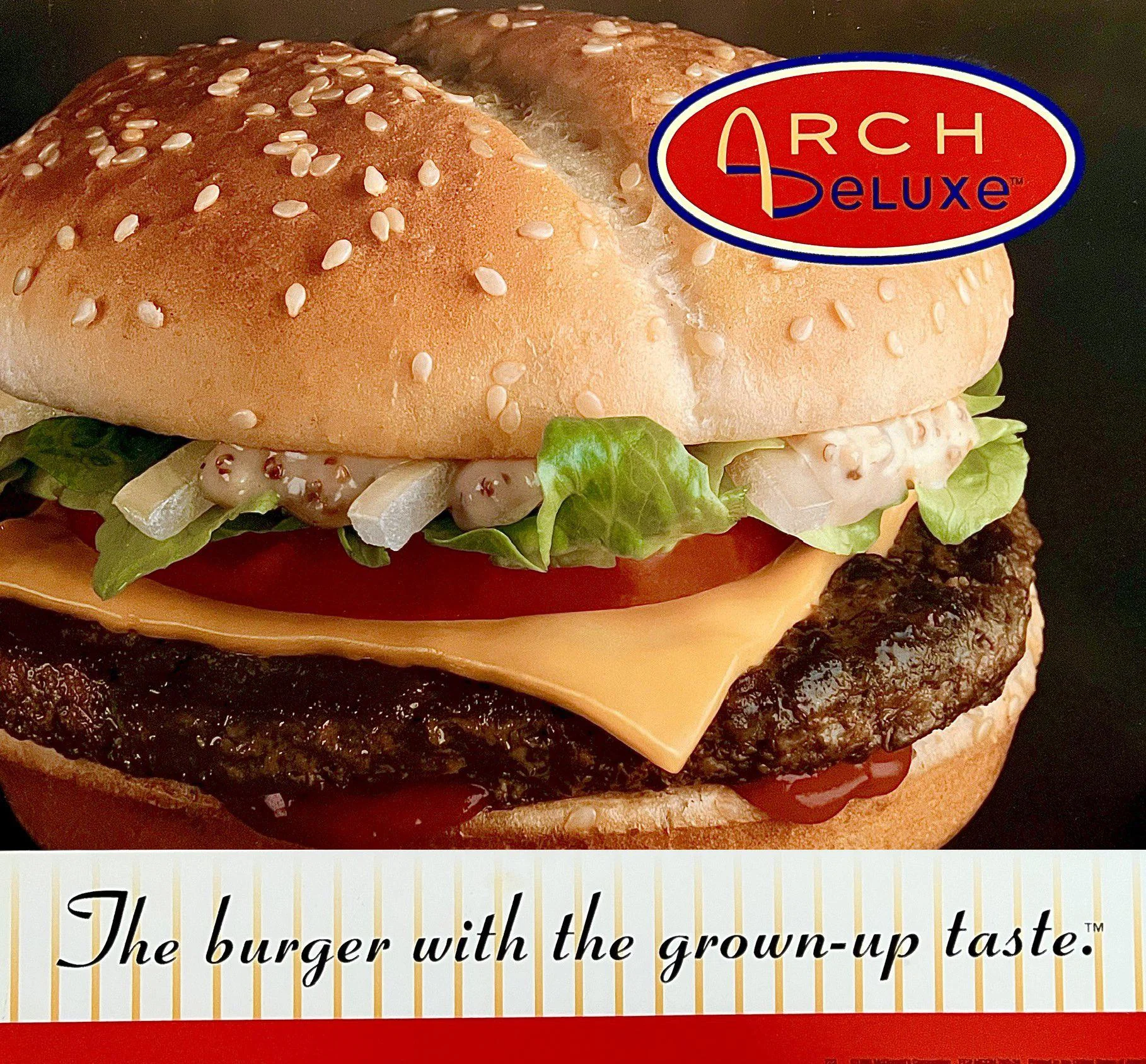 A close-up of a sesame seed hamburger bun sandwiching lettuce, tomato, onions, cheddar cheese, ketchup, and a beef patty. The top right corner has an "Arch Deluxe" logo, with the tagline "The burger with the grown-up taste.
