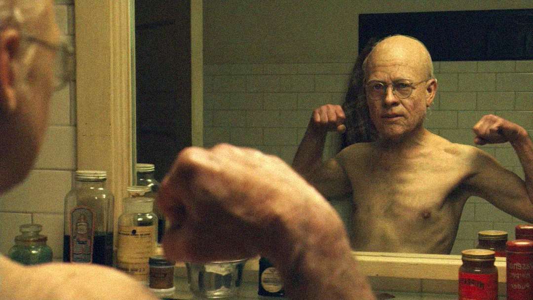 An elderly, bald man with glasses stands shirtless in front of a bathroom mirror, flexing his arms. Various bottles and toiletry items are visible on the sink and shelves.