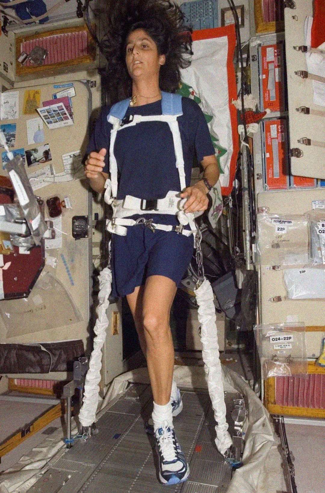 An astronaut with long hair jogs on a treadmill in a spacecraft, wearing a harness to stay anchored. Equipment, storage bags, and control panels line the walls around her.