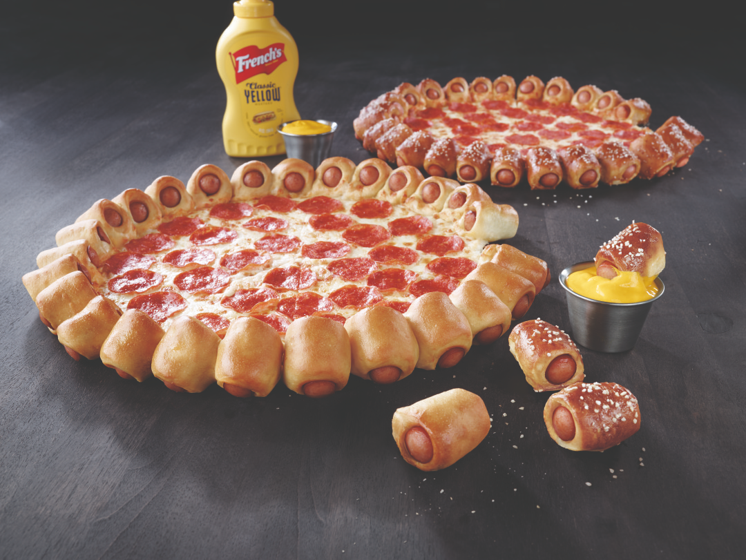 A pepperoni pizza with a crust made of mini hot dogs in dough, served with mustard and cheese dip. A bottle of French's yellow mustard and another similar pizza are in the background.