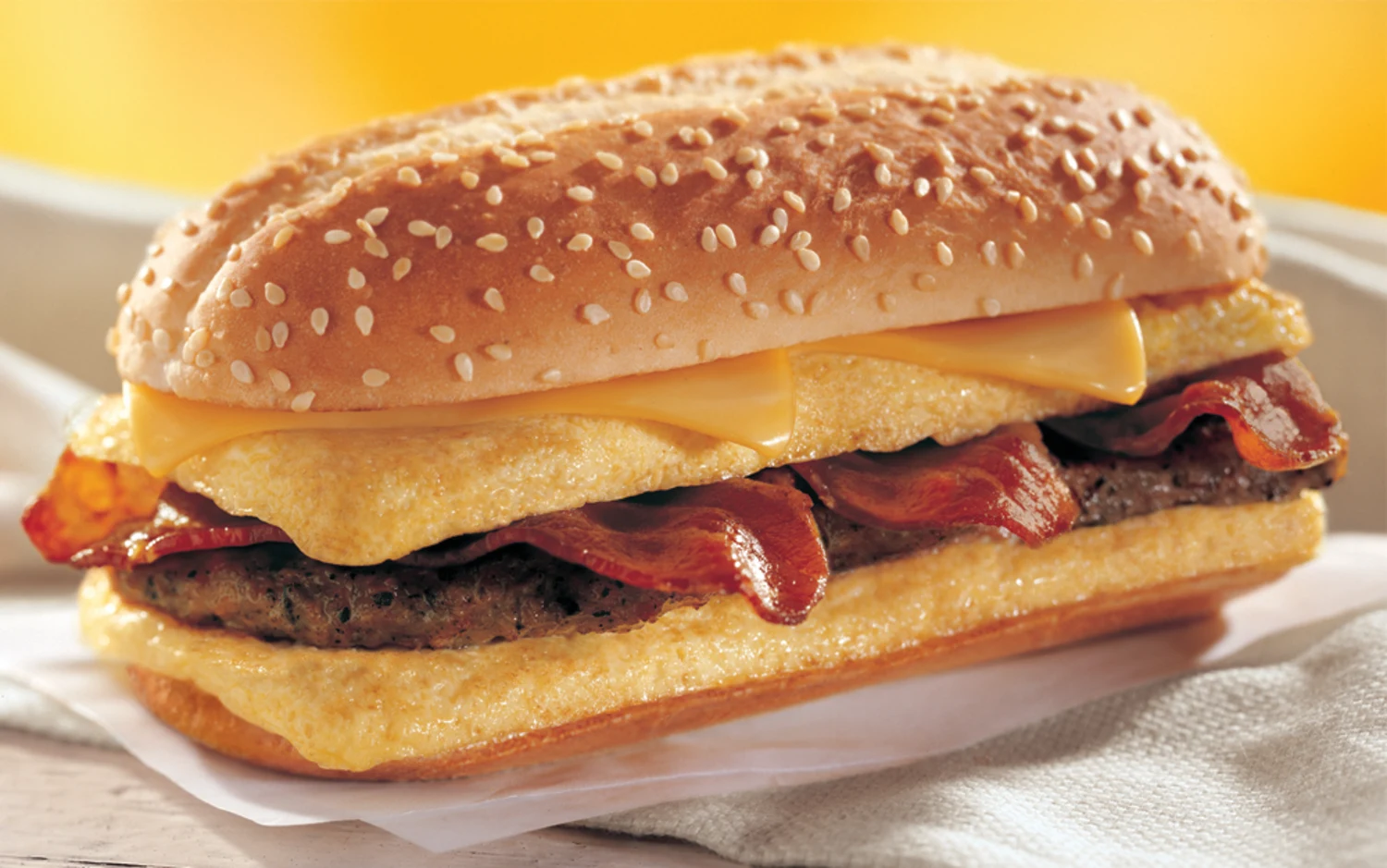 A breakfast sandwich with a sesame seed hoagie bun, folded egg, crispy bacon strips, a sausage patty, and two slices of melted cheese, placed on white paper with a soft yellow background.