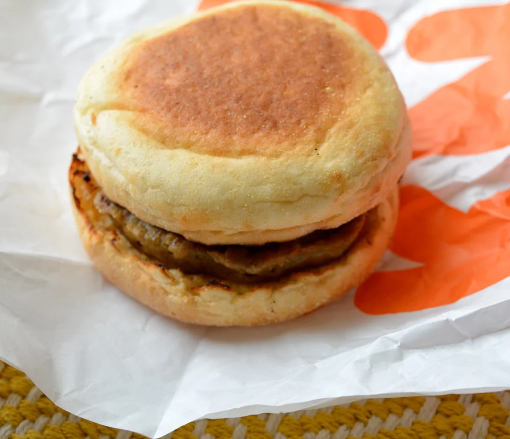 A sausage patty breakfast sandwich on an English muffin sits on white and orange fast food wrapper.