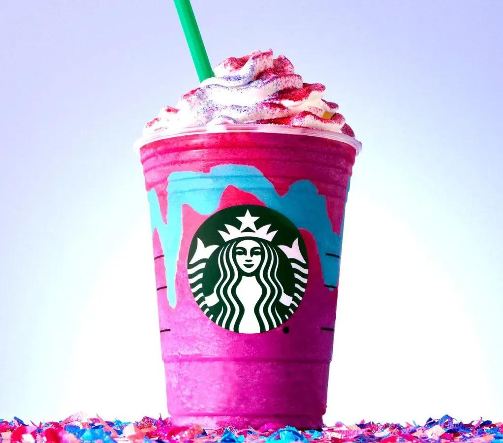 A vibrant Starbucks cup filled with a colorful, swirled pink and blue blended drink, topped with whipped cream and sprinkles. A green straw sticks out of the cup, which is surrounded by scattered confetti.