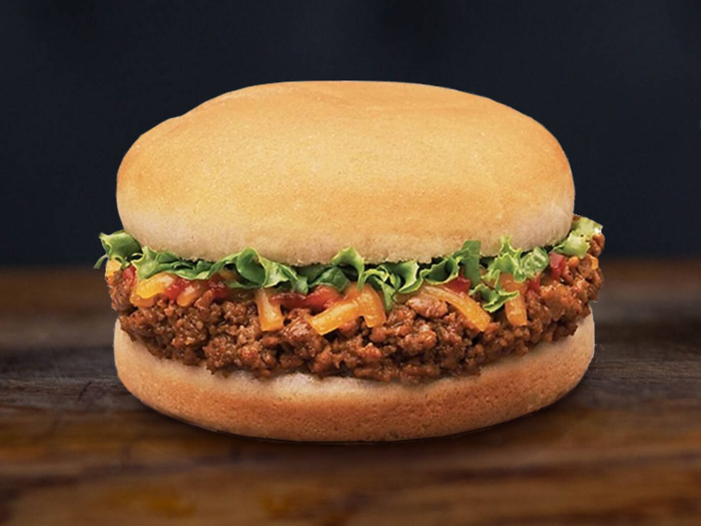 A sloppy joe sandwich with ground beef, shredded cheese, lettuce, and sauce on a soft bun, placed on a wooden surface with a dark background.
