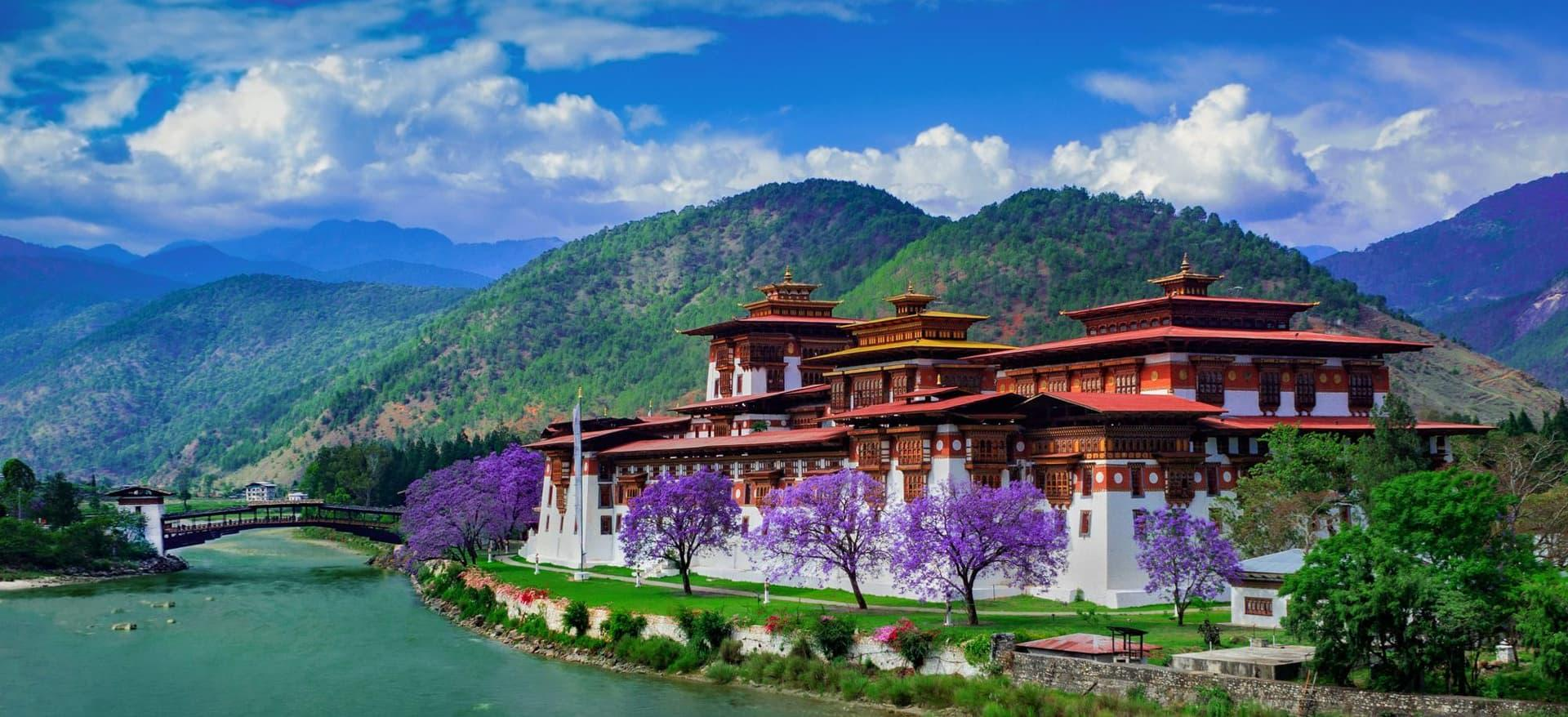 A large traditional Bhutanese fortress with red and gold roofs stands by a river, surrounded by blooming purple trees and green mountains under a blue sky with scattered clouds.