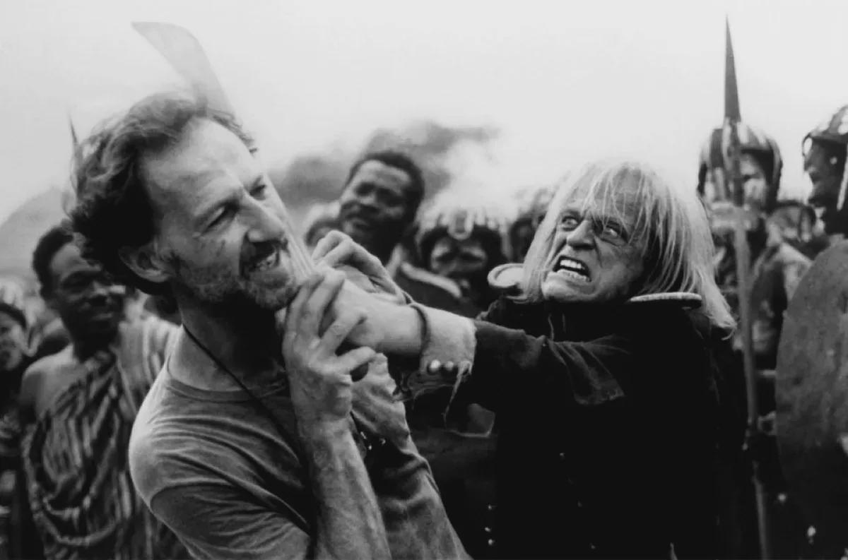 A black-and-white photo shows two men in a struggle; one grabs the other’s neck with a fierce expression. Several onlookers and people in historical costumes stand in the background.