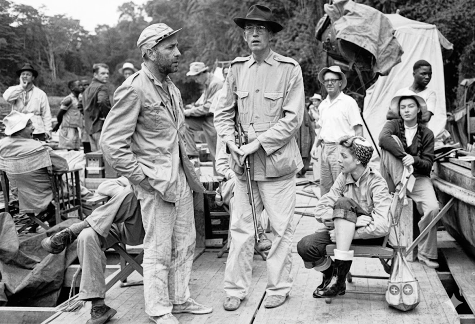 A black-and-white photo shows a film crew and actors on a crowded wooden set, outdoors. Two men in front wear rugged clothing and hats; one holds a rifle. Equipment and people surround them amid a jungle backdrop.
