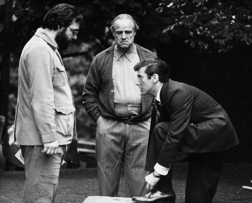 Three men are gathered outdoors; one in a casual jacket looks down, another in a cardigan stands with hands in pockets, and a third man in a suit polishes his shoe on a bench, while the other two watch.