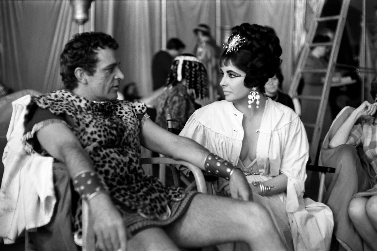 A man and woman in ancient-style costumes sit and talk backstage on a film set; the man wears a leopard-print tunic, and the woman has an elaborate hairstyle, large earrings, and a bejeweled headpiece.