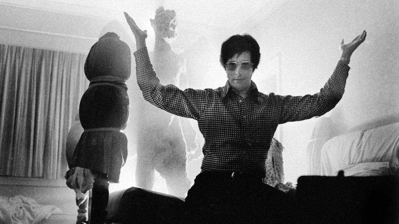A person wearing glasses raises their arms while sitting in a dimly lit room, with a hazy, shadowy figure and dramatic lighting in the background. A wrapped object stands to the left and a sofa is visible on the right.