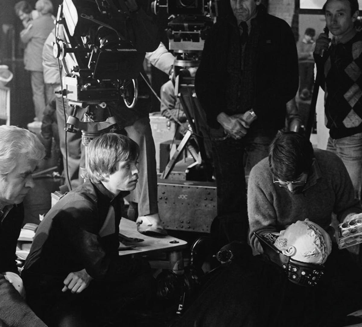 Behind-the-scenes black-and-white photo shows crew members and actors on a film set. One actor lies down with a prosthetic mask, surrounded by people, cameras, and filming equipment.