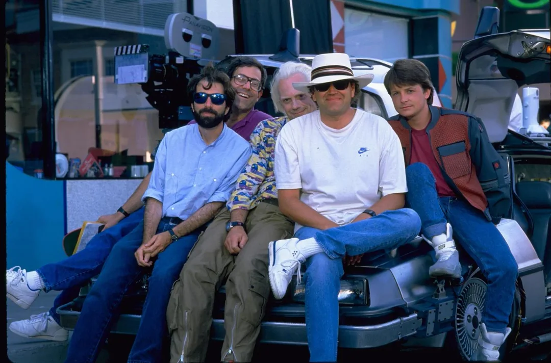Four men sit on the hood of a DeLorean car, with a film camera behind them on set. Two are wearing sunglasses, and one is dressed in a red jacket, resembling a character from "Back to the Future.