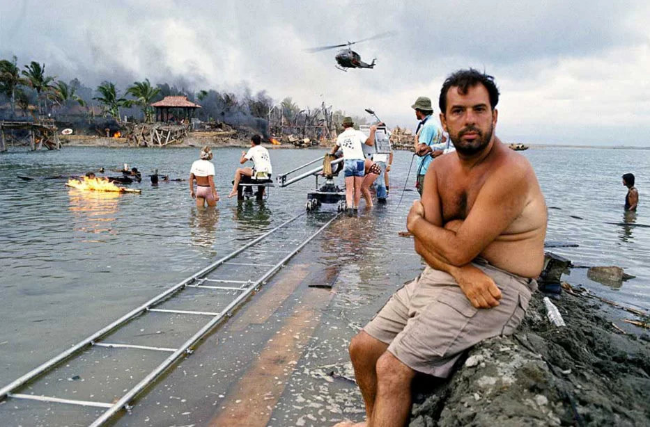 A shirtless man sits on muddy ground near the shore, arms crossed, as a film crew works in shallow water with camera equipment. In the background, a helicopter flies above burning buildings and palm trees.