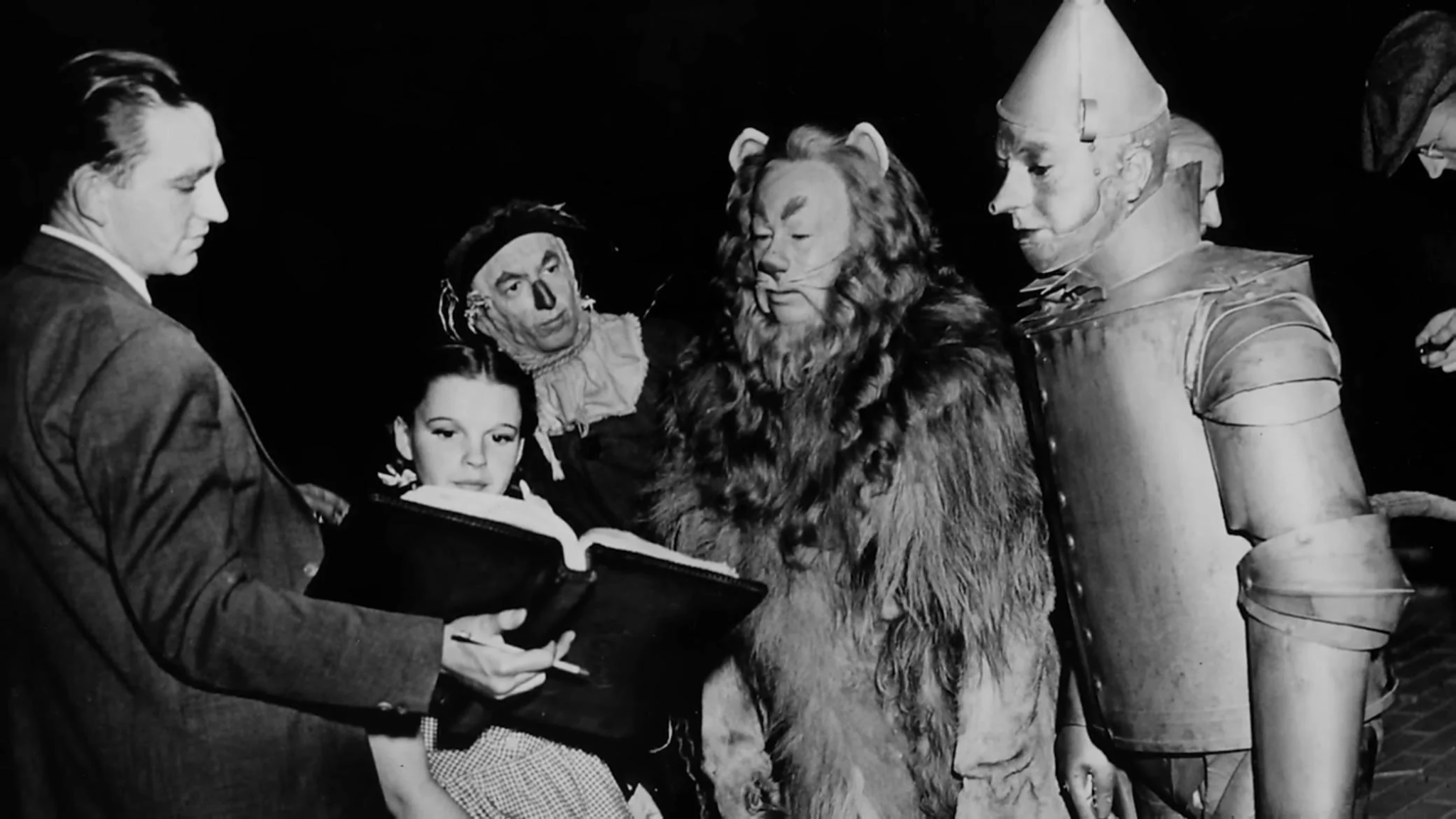 A black-and-white photo shows four people in elaborate costumes, including a lion, a tin man, and a scarecrow, gathered around a seated young woman and a man holding an open book, possibly discussing a script.