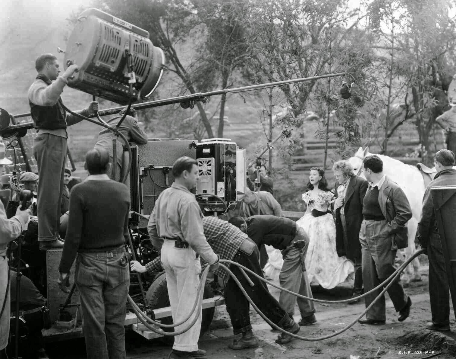 A black-and-white scene shows a film crew operating cameras, lights, and a boom mic on an outdoor set, while an actress in a long dress poses with a man and a white horse in the background among trees.
