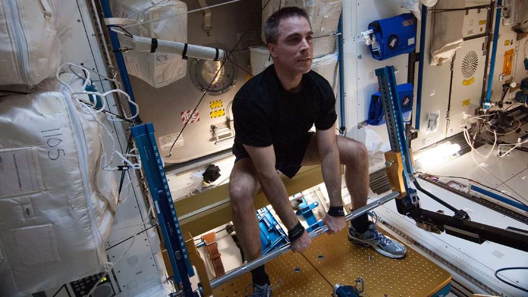 An astronaut in a black shirt and shorts performs a weightlifting exercise on specialized equipment inside a space station, surrounded by various instruments and storage containers.