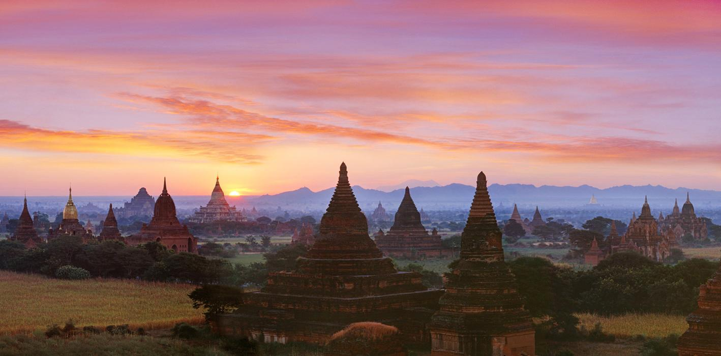 Ancient temples and pagodas of Bagan, Myanmar, are silhouetted against a colorful sunrise sky, with fields and distant mountains in the background.