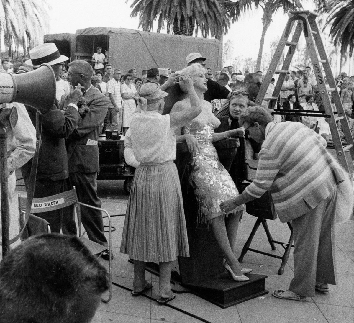 A glamorous woman in a sparkling dress sits on a platform surrounded by crew members adjusting her outfit and makeup on a busy outdoor film set; palm trees and equipment are visible in the background.