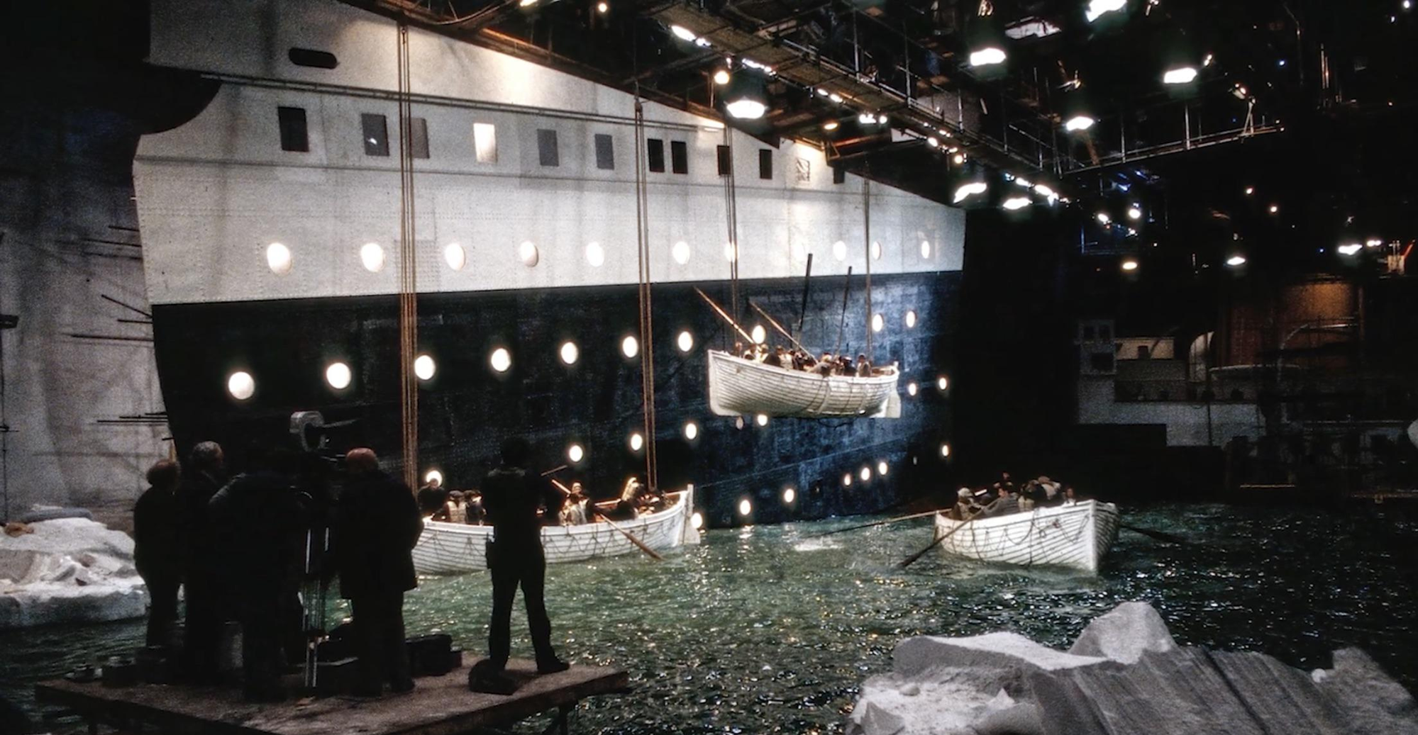 A film set showing lifeboats with actors being lowered from a large replica ship’s side into a water tank, surrounded by crew, cameras, and lighting equipment, recreating a Titanic scene indoors.