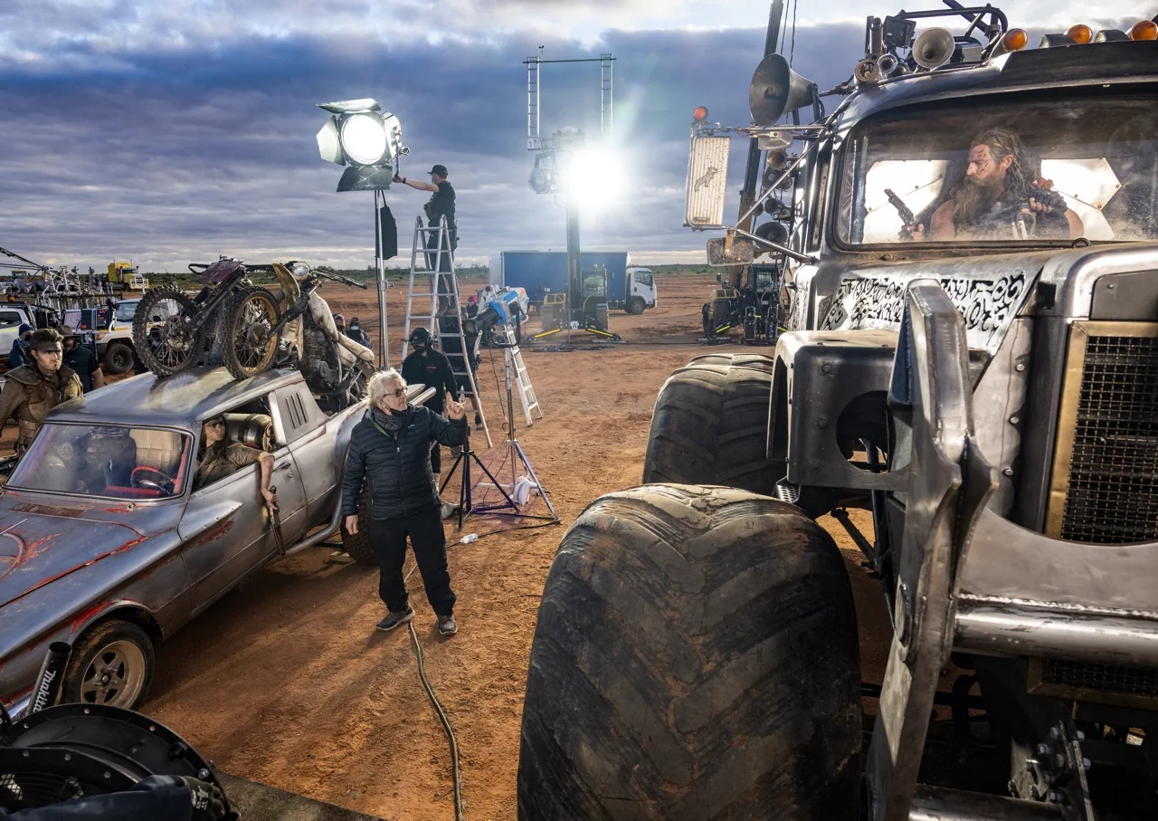 On a film set, crew members adjust lighting and equipment around rugged vehicles in a desert setting. One person stands between a silver car and a large truck with an actor inside. Bright lights and cameras are visible in the background.