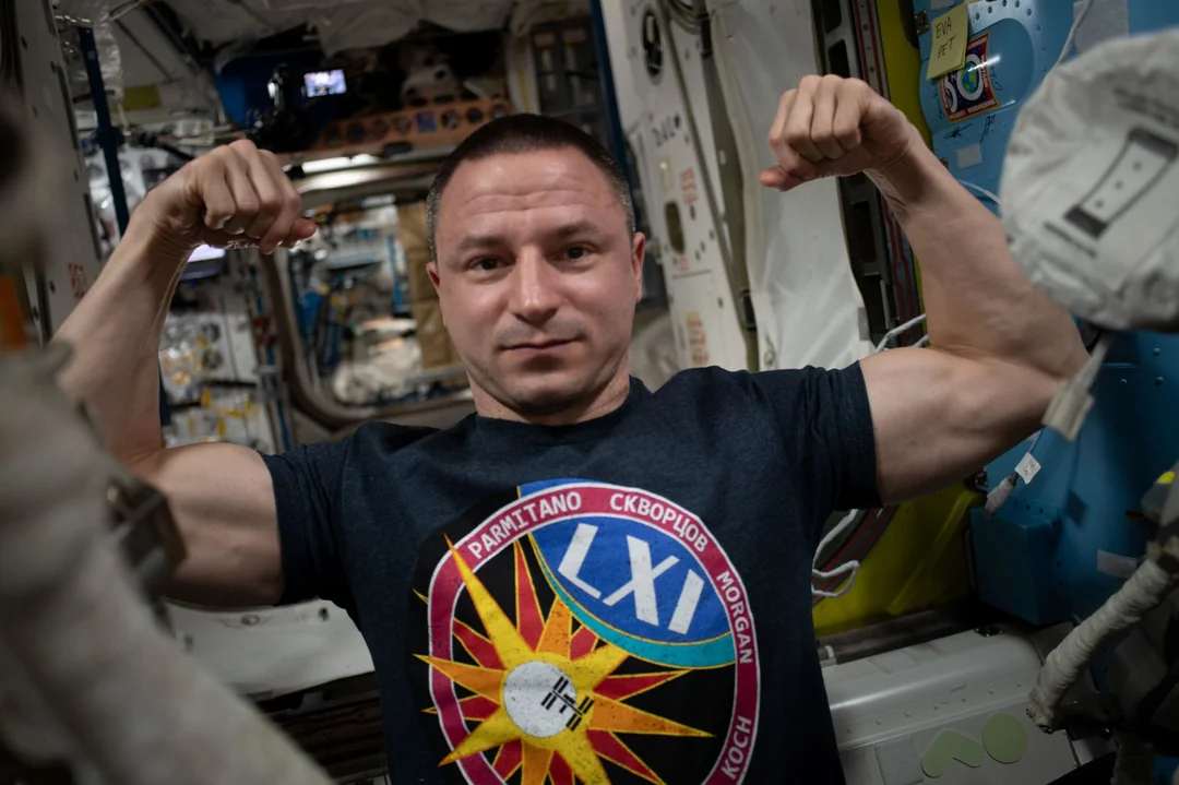 A person in a dark t-shirt flexes their muscles and smiles inside a space station, surrounded by equipment and cables in a compact, well-lit environment.