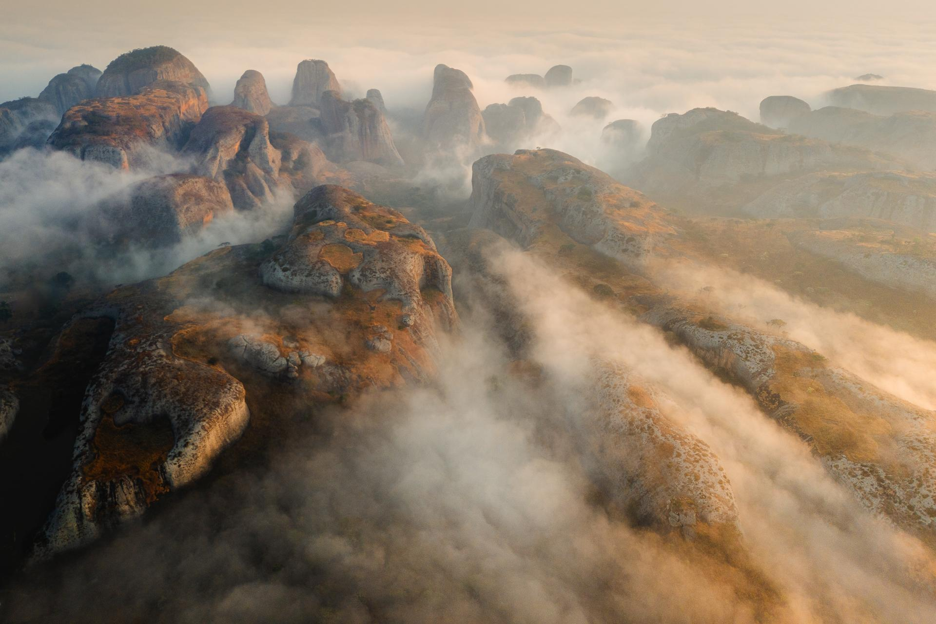 Aerial view of rocky cliffs and large stone formations rising above a layer of mist and clouds, bathed in warm golden light, creating a dramatic and ethereal landscape.