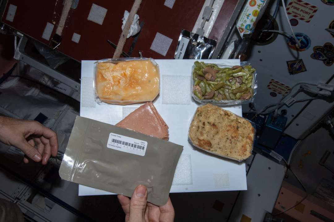 A person holds a vacuum-sealed food pouch above a tray with packaged foods, including cheese, beans, bread, and meat, inside a space station with equipment and labels visible in the background.