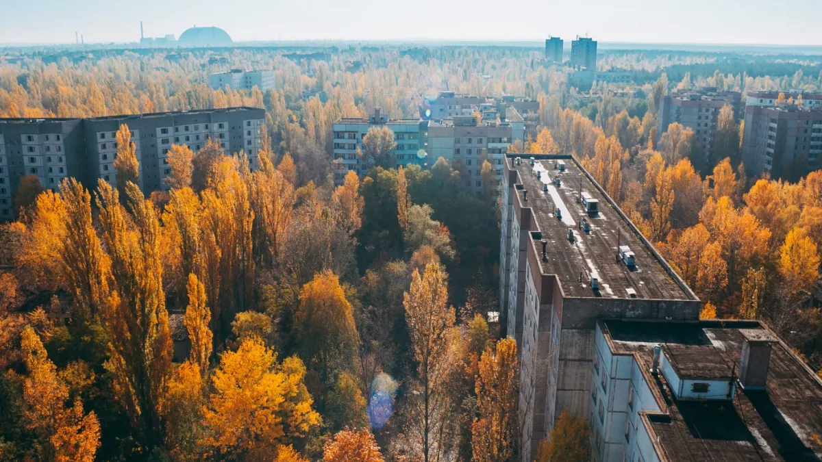 Aerial view of abandoned apartment buildings surrounded by dense autumn trees with yellow-orange leaves; distant structures and a large dome are visible on the horizon under a hazy sky.