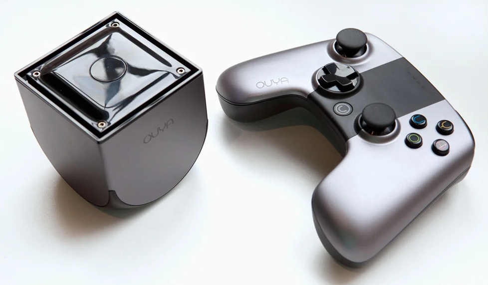 A silver and black Ouya game console sits next to its matching wireless controller with two joysticks, a directional pad, and colored buttons, all on a white surface.