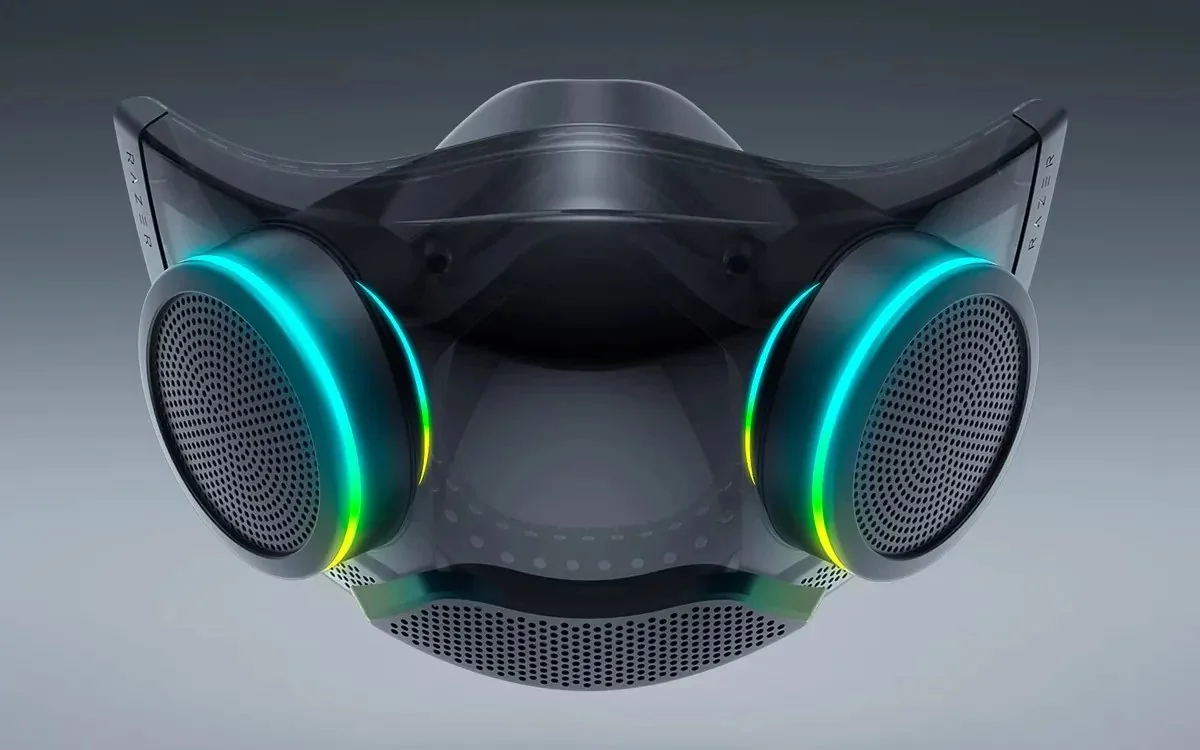 A futuristic black face mask with two large circular filters on each side, both illuminated with blue and green LED rings, and a transparent section in the center. The background is a gradient of gray tones.