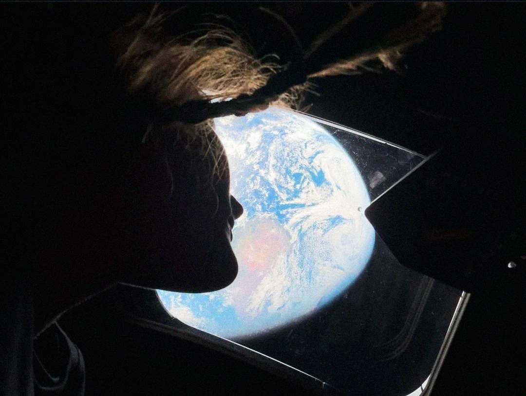 A person with loose hair gazes out of a spacecraft window at Earth, which appears bright and blue against the darkness of space.