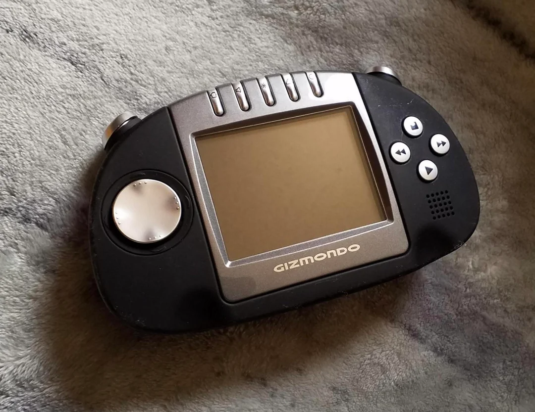 A handheld Gizmondo game console with a large central screen, directional pad on the left, and various control buttons on the right, resting on a soft, gray textured surface.