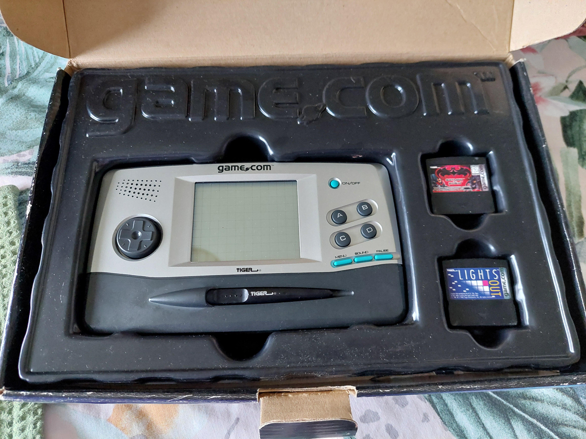 An open box containing a vintage Tiger game.com handheld console, a stylus, and two game cartridges, all neatly placed in black plastic packaging.
