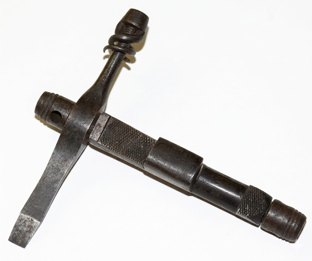 A vintage metal multi-tool with a T-shaped design, featuring a flathead screwdriver on one end and knurled grips along the handle, set against a plain white background.