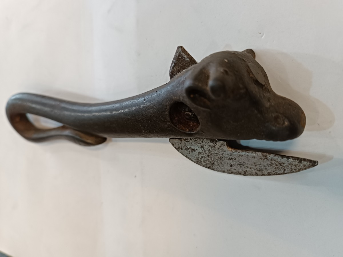 A vintage metal can opener shaped like an animal's head, possibly a dog or horse, with a curved handle and a rusty blade, placed on a white surface.