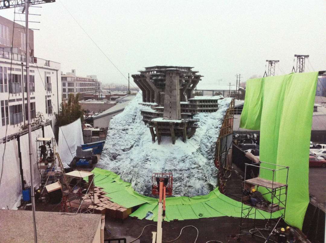 A film set featuring a large, detailed model of a ruined bridge on a snowy hill, surrounded by green screens and production equipment, with buildings and scaffolding in the background.