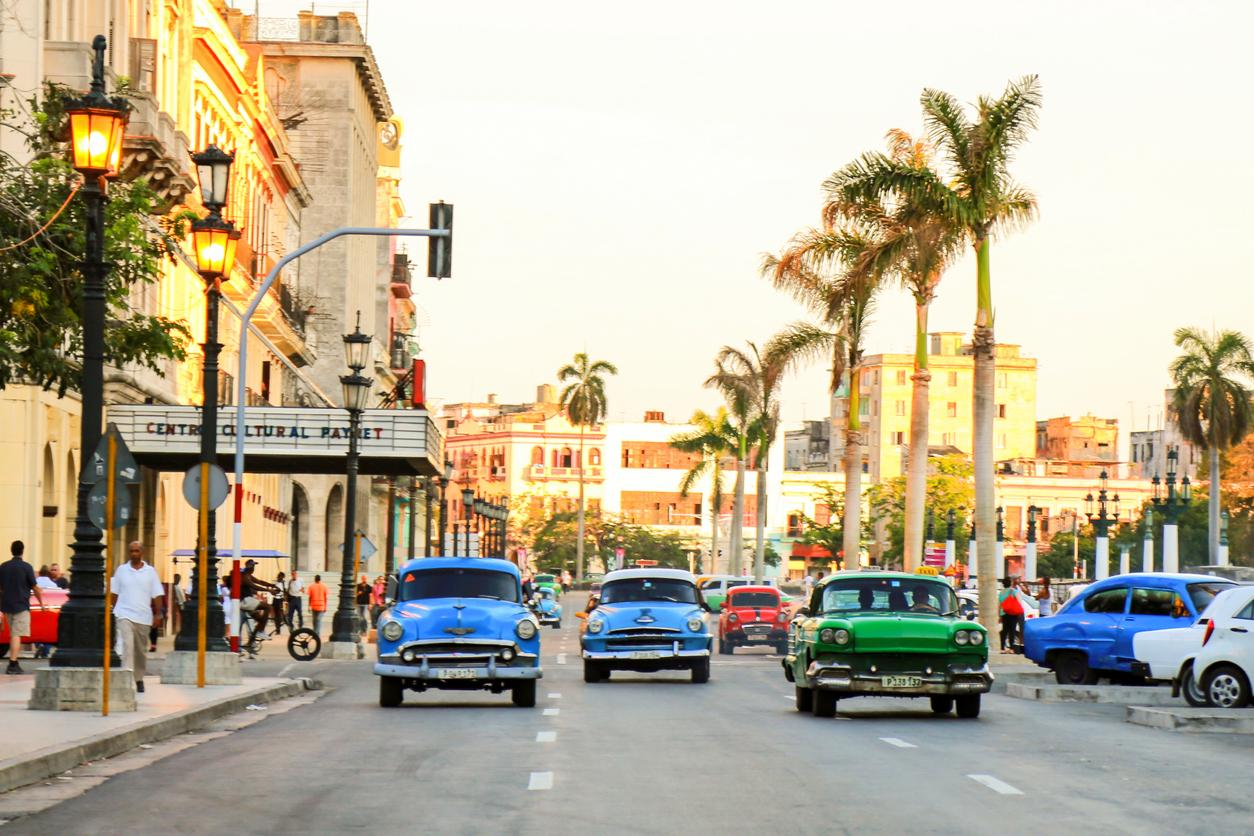 Colorful vintage cars drive down a street lined with palm trees and historic buildings at sunset in Havana, Cuba. People walk on the sidewalks, and the Centro Cultural Payret is visible on the left.