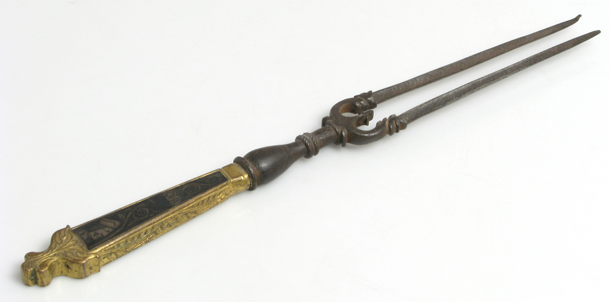 An antique meat fork with two long, rusted prongs and an ornate handle featuring gold and black decorative patterns, displayed on a white background.