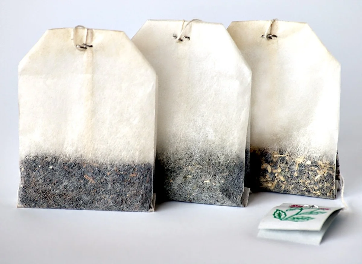 Three paper tea bags filled with loose tea leaves are standing upright on a white surface. Each bag has a string with a paper tag attached, and they are arranged closely together.