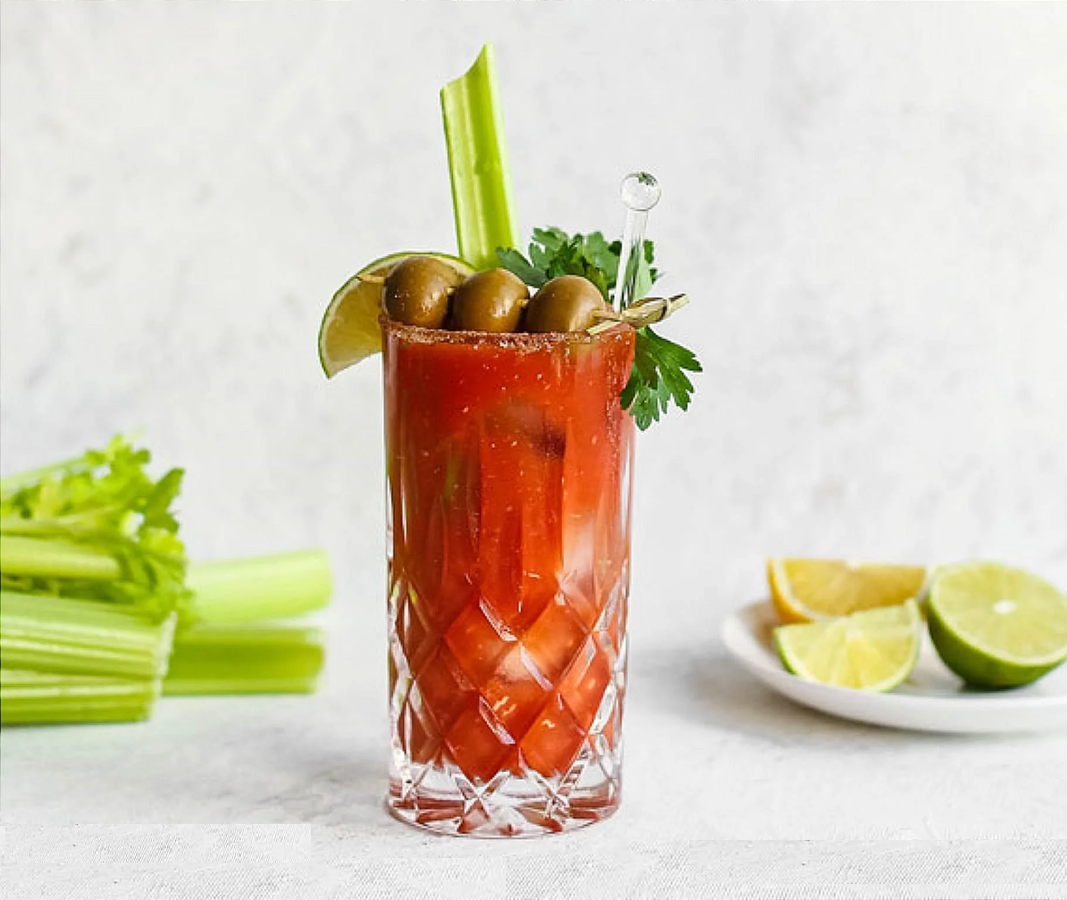 A glass of Bloody Mary garnished with green olives, a celery stick, a lime wedge, and parsley, sits on a white surface. Celery stalks and a plate with lime and lemon wedges are in the background.